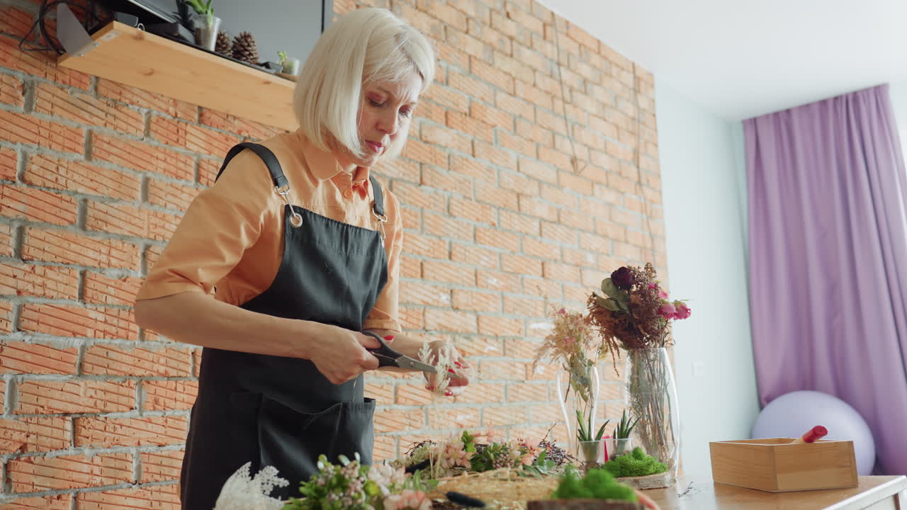 Blonde woman in black apron focused on arranging flowers and dried plants on wooden table against brick wall, demonstrating floral craft with natural materials in cozy indoor creative workspace