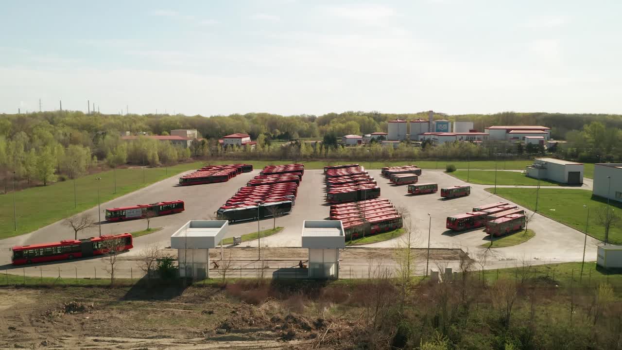Slowly flying over a parking lot for public transportation buses. One red  bus is driving in and parking. Industrial background on transportation theme. The camera is slowly descending.
