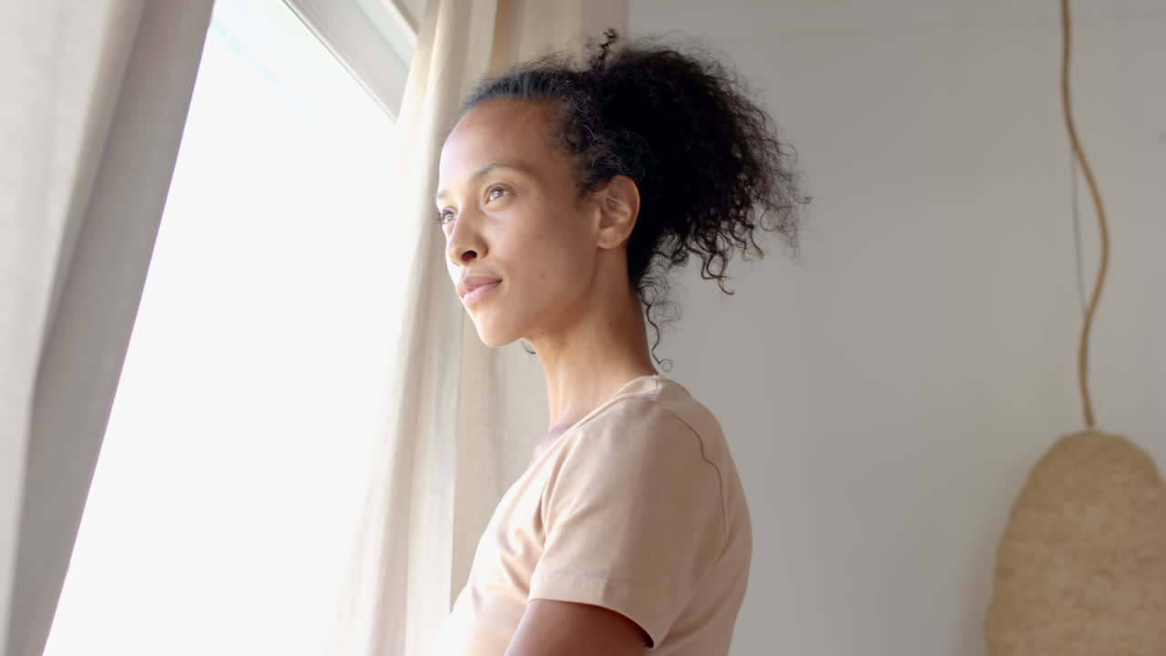 Woman standing by window at home, gazing outside with thoughtful expression