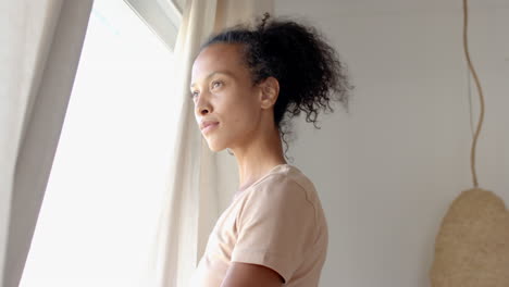 woman standing by window at home, gazing outside with thoughtful expression