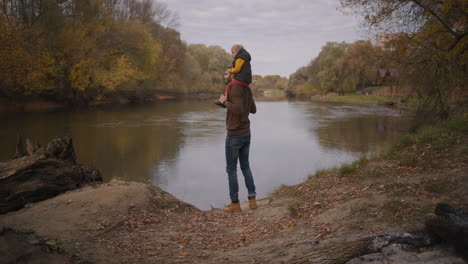 young father is showing nature and autumn landscape to his little son holding child on shoulders standing on lake coast back view of human