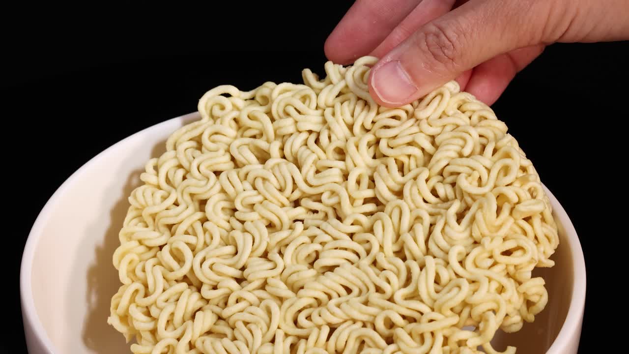 A hand places a round block of uncooked instant noodles into a clean white bowl, under bright studio lighting with a black backdrop and smooth camera movement