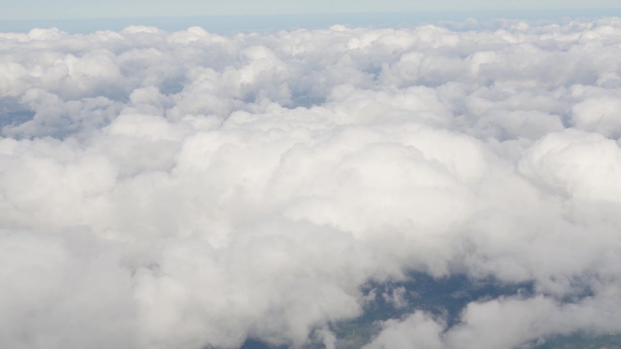 Dense clouds fill the frame seen from an aircraft window. The view shows various cloud layers and shadows. Ground features are partially visible beneath the clouds.