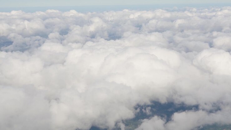 Dense clouds fill the frame seen from an aircraft window. The view shows various cloud layers and shadows. Ground features are partially visible beneath the clouds.