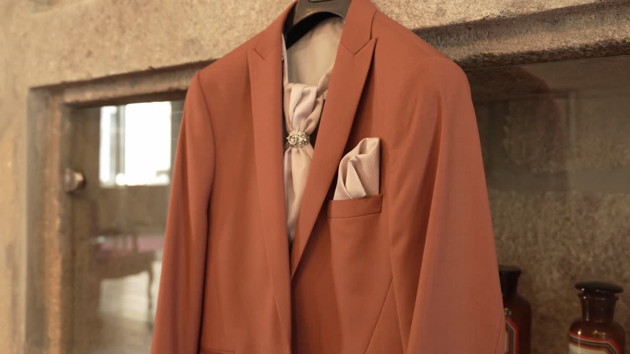 Close up of an elegant terracotta suit with cream tie and pocket square hanging on a stone wall, ready for a wedding
