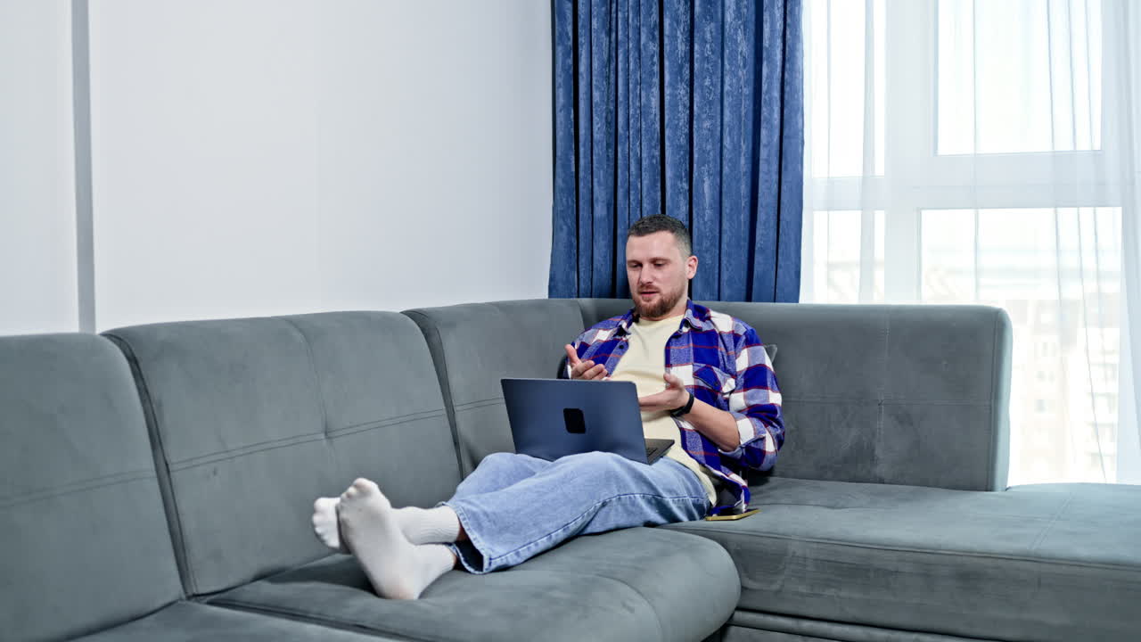 Man sitting on couch in living room working on laptop during day while wearing casual clothes and talking