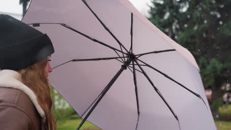 student holding umbrella, wearing black knit cap and brown shearling jacket, walking calmly during cold overcast day with light snowfall, immersed in peaceful winter landscape