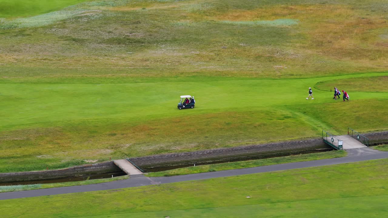 Golfers and caddies walk across bridge on lush fairway, wide aerial drone shot, daylight