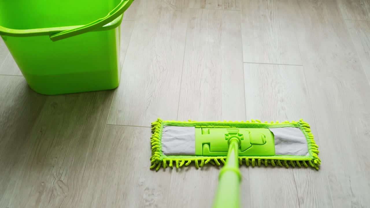 Green mop and bucket on wooden floor at home. Housewife cleaning the parquet floor with a modern microfiber brush. Daily routine.