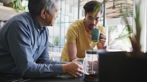 Man-talking-to-young-son-drinking-coffee-from-mug-while-leaning-on-kitchen-counter-at-home