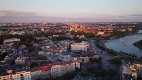 Aerial-backwards-shot-over-beautiful-lighting-city-of-Krakow-with-Vistula-River-and-Wawel-Royal-Castle-and-cruising-boats-at-sunset