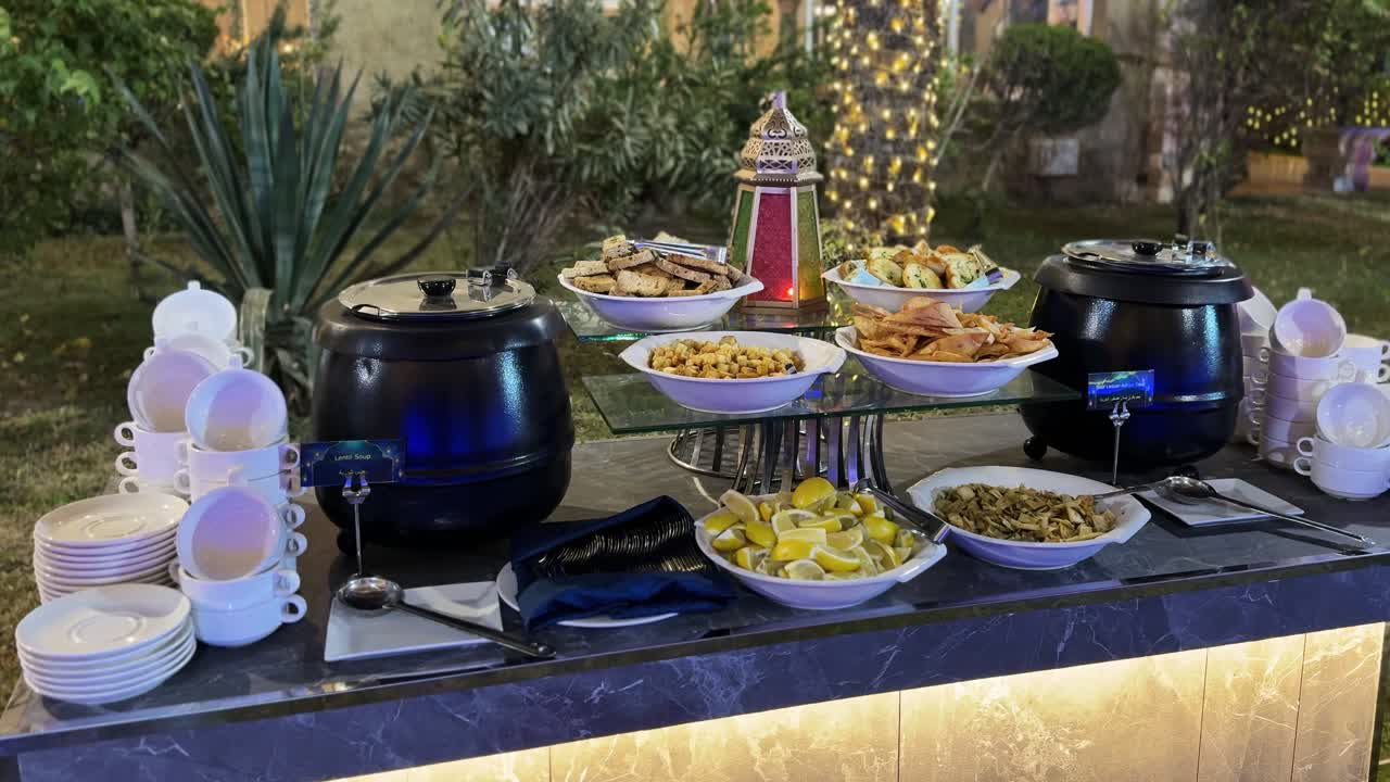 Esposizione di catering serale in giardino con bollitori neri per zuppa etichettati "zuppa di lenticchie", tazze impilate, spicchi di limone, crostini e centrotavola a lanterna sotto calde luci festive.