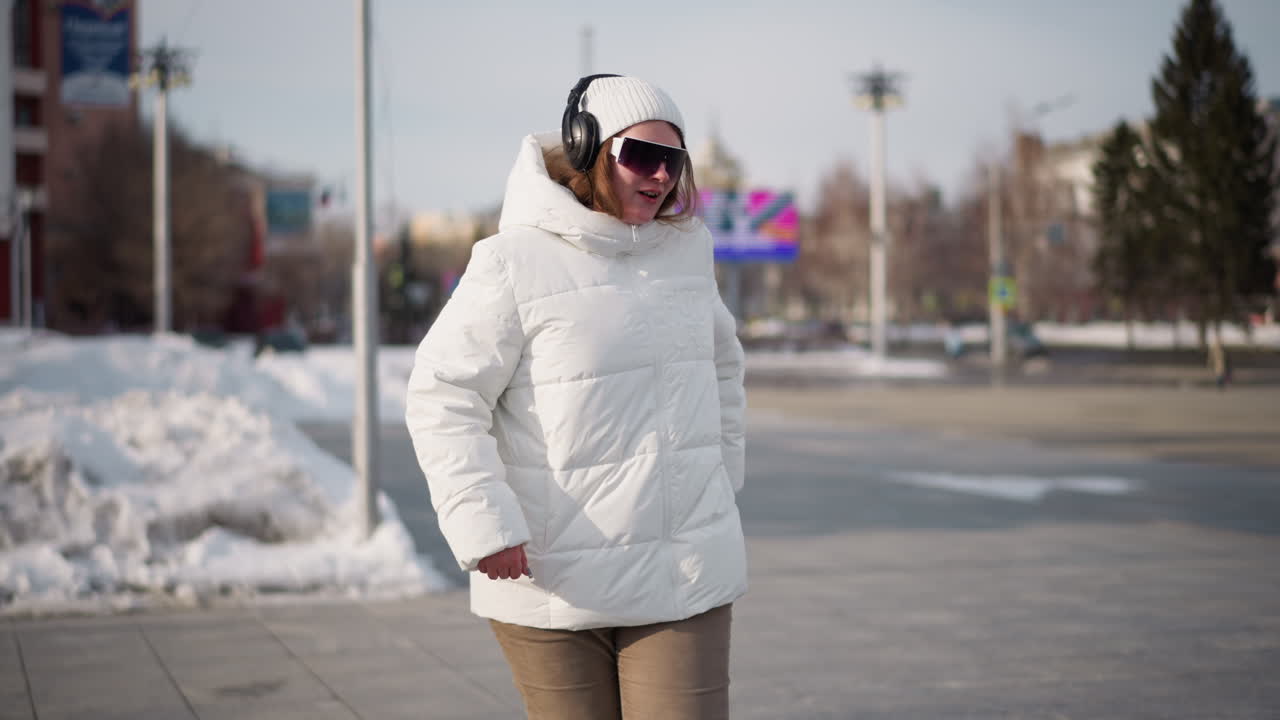 Young lady wearing white winter coat, beanie, black headset and white goggles performs dance moves on tiled snowy plaza under bright daylight with cars passing on road and trees framing view