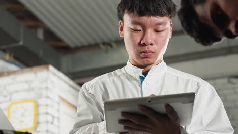 chinese student in lab coat taking notes with tablet while partially visible man observes, industrial mechanical workshop setting with tools and equipment in background