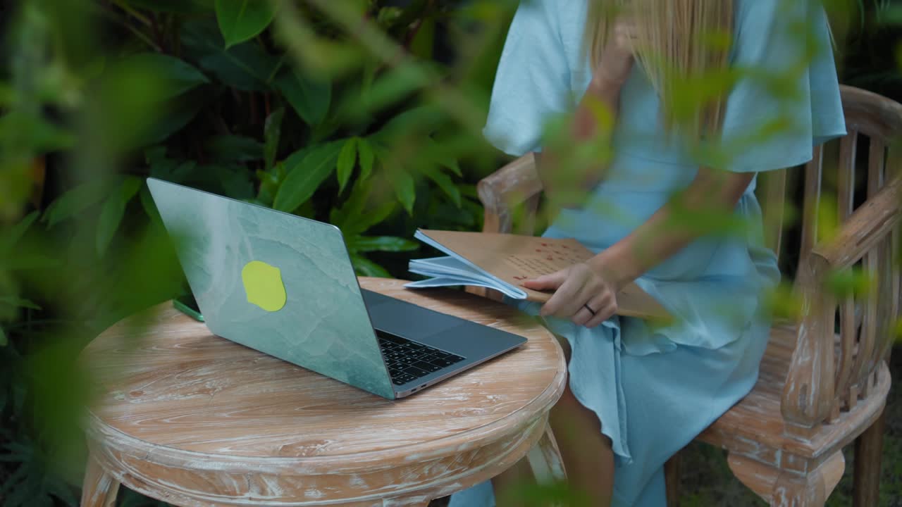 una chica en un vestido azul remotamente en línea trabajando detrás de una computadora portátil y mirando a la pantalla el patio trasero con plantas verdes