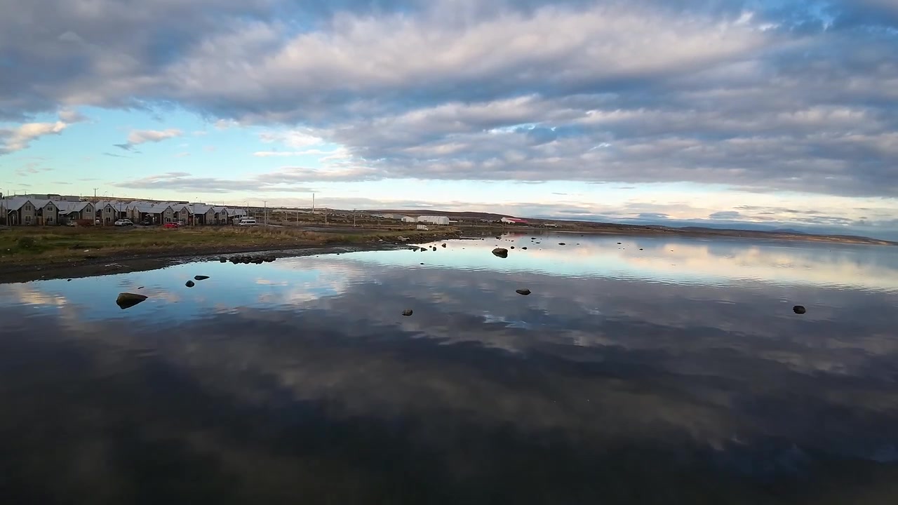Rock strewn shoreline of Última Esperanza Fjord reflecting a cloudy sky, with a new residential neighborhood along the water in Puerto Natales, Chile, FPV aerial