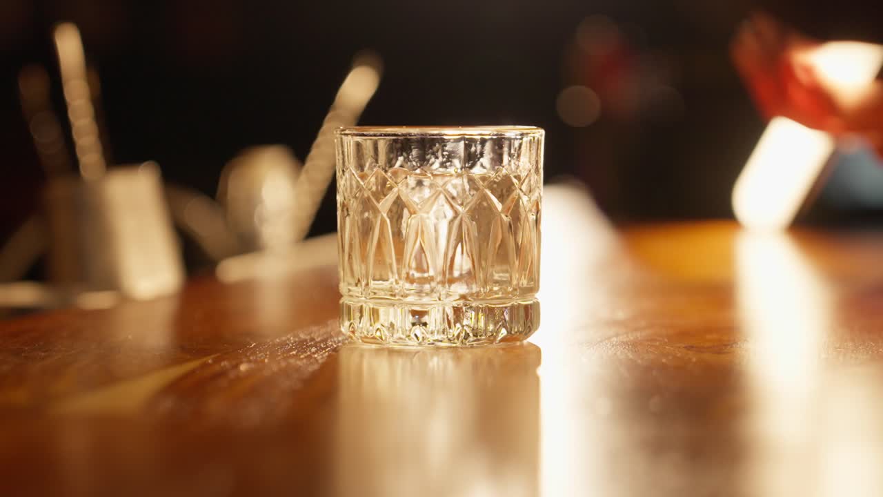 Primo piano di un barista che usa delle pinze per posizionare un cubetto di ghiaccio trasparente scolpito a mano in un bicchiere rocks di cristallo, facendolo ruotare per raffreddare il bicchiere in un bar.