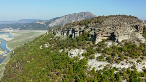 An-orbiting-aerial-view-of-a-mountain-top-surrounded-by-green-trees,-with-a-river-and-blue-sky-in-the-background