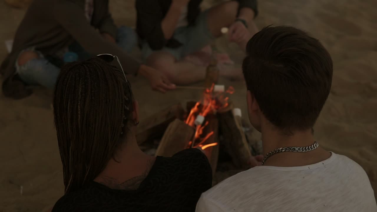 해변에서 불  ⁇ 에 앉아 마시멜로를  ⁇ 고 있는 젊은 친구들의 그룹. 느린 움직임으로 촬영