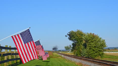view of a steam passenger train approaching blowing smoke and steam with american flags waving in the wind on the fence