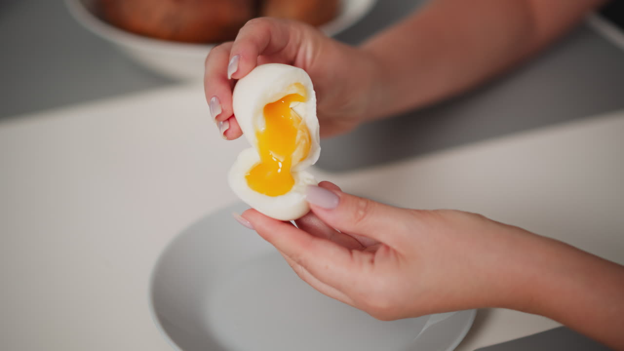 Close up of person holding soft-boiled egg with runny yolk partially turned into other half of white shell over plate, showing texture and gentle handling of fragile egg during food preparation