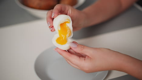 close up of person holding soft-boiled egg with runny yolk partially turned into other half of white shell over plate, showing texture and gentle handling of fragile egg during food preparation