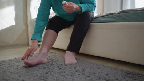 old woman in green applying ointment to her hand while sitting on the edge of the bed. one sock on, the other off, as she takes care of herself in a cozy, peaceful bedroom setting with soft light