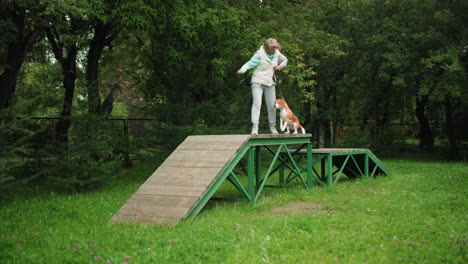 dog trainer running through obstacle course with her dog reaching finishing line rewarding with food in grassy natural environment surrounded by trees during outdoor training session bonding with pet