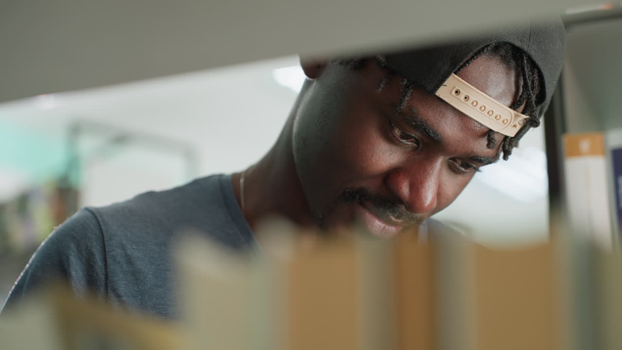 Close up of young man wearing cap and casual shirt standing near bookshelf in library, focusing intently on books with slight smile, surrounded by colorful book spines and bright indoor lighting