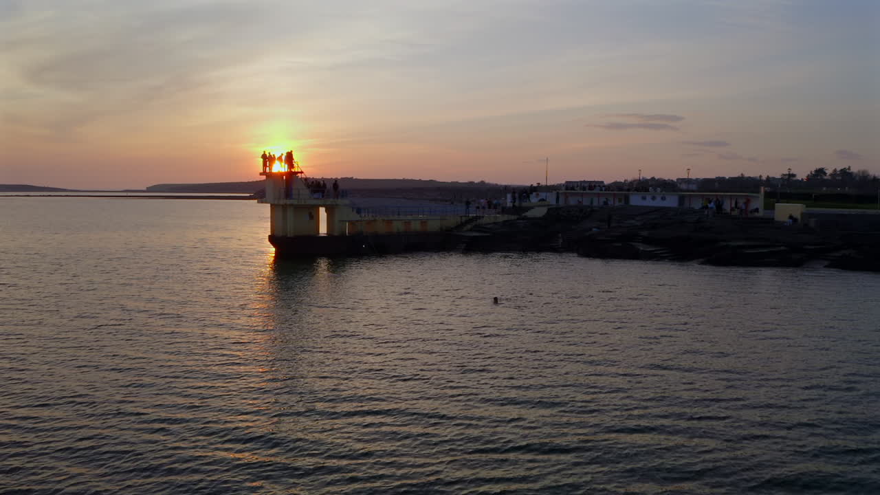 Diving tower against soft orange sky and mirrored water surface, backlit silhouette of people standing at edge, Galway Ireland
