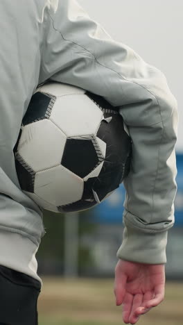 back view of a person standing with a soccer ball tucked under their right arm, wearing a gray jacket and blue pants, with a blurred view of a grassy field and a blue building