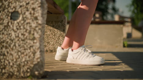 leg view of young white lady sitting on bench wearing white canvas shoes with shadow cast on leg, blurred background featuring greenery and stone bench base