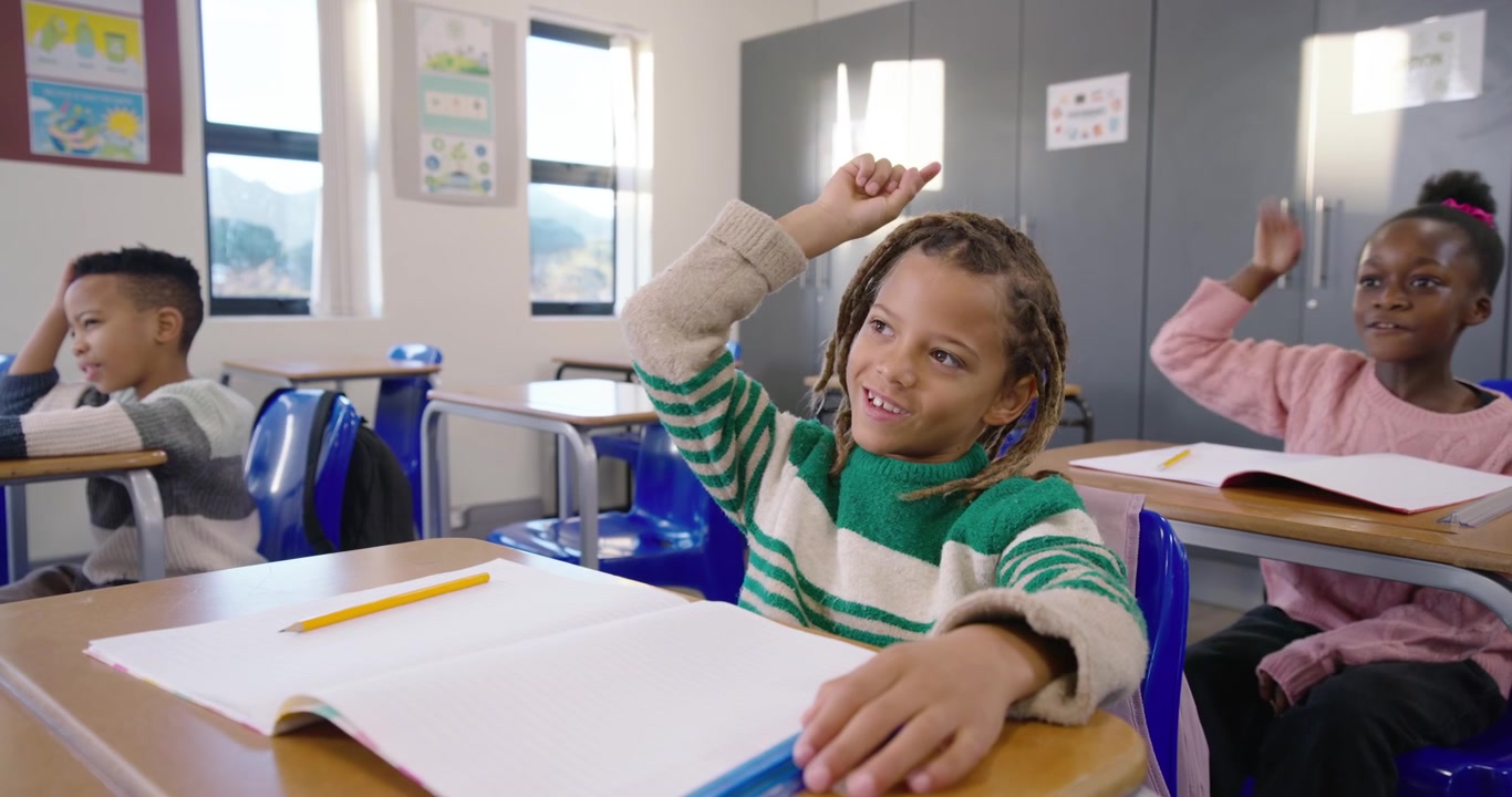 African American girl in green top answering teacher cue, raising hand and writing notebook at desk