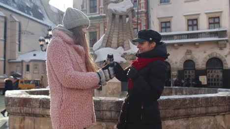 Two-smiling-women-tourists-traveling-together,-drinking-hot-tea,-coffee-from-thermos-on-city-street