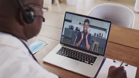 african american male doctor wearing phone headset taking notes while having a video call on laptop