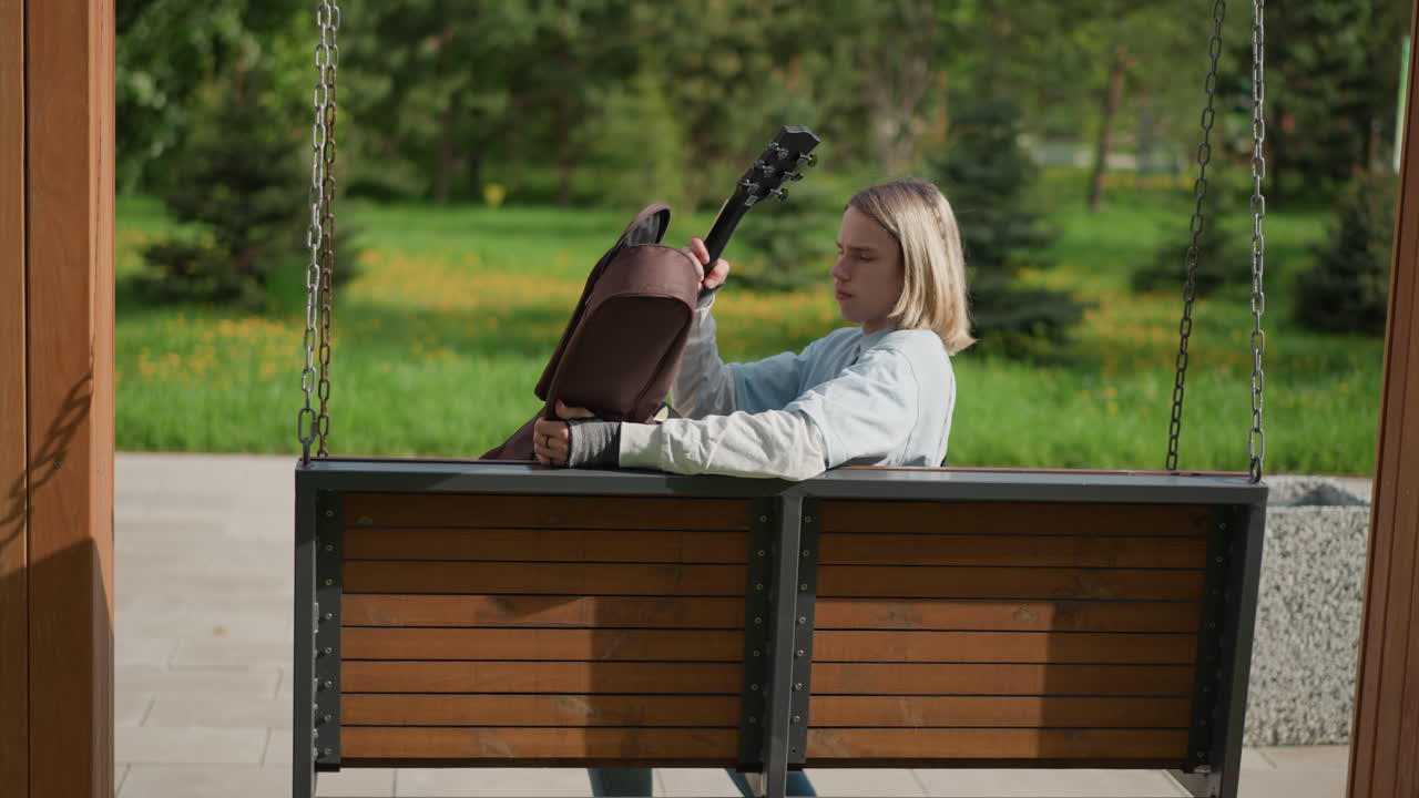 Woman guitarist on porch swing opening guitar case, lifting acoustic instrument, tuning strings, then strumming gentle melody. Sunlit pergola, wooden bench with chain supports, green park background