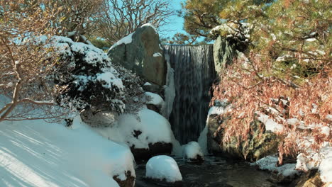 Langsamer-Schwenk-Rechts-Vom-Kleinen-Wasserfall-Im-Winter-Mit-Schneebedeckten-Pflanzen