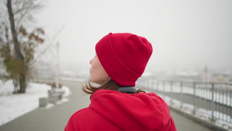 vista de cerca de la mujer con gorra roja y chaqueta mirando hacia arriba con auriculares alrededor del cuello, rodeada de un entorno de polvo de nieve, barandilla de hierro y paisaje urbano borroso en una atmósfera de invierno nebulosa