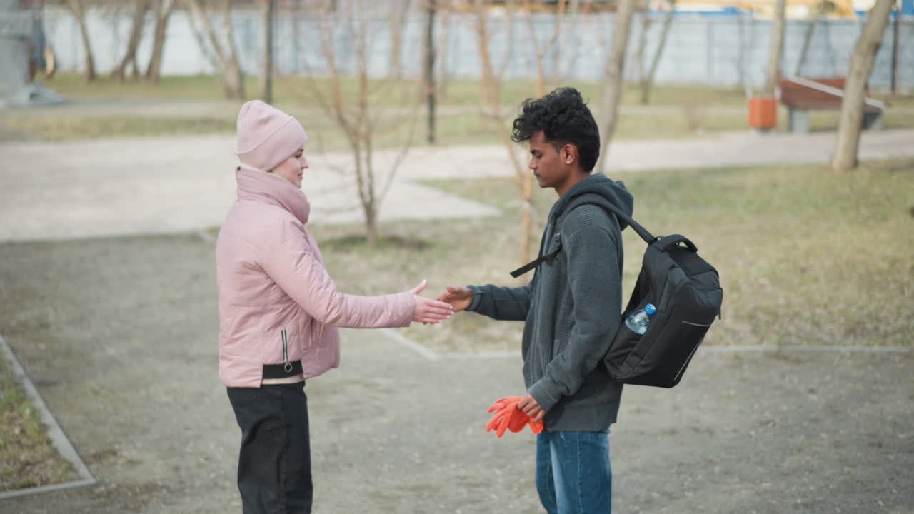 Pria berkulit hitam mengenakan hoodie abu-abu gelap membawa ransel hitam memegang sarung tangan oranye, berdiri berhadapan dengan wanita berjaket merah muda dan topi kupluk di luar ruangan di taman saat awal musim semi, dikelilingi oleh pohon-pohon tanpa daun.