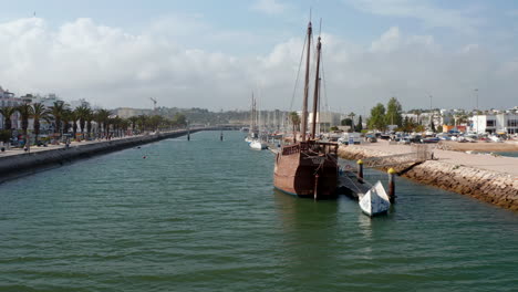 Portuguese-wooden-caravela-moored-in-harbor-town-of-Lagos-Algarve.-Aerial-drone-flying-forward-towards-sailing-boat-in-port-canal,-day