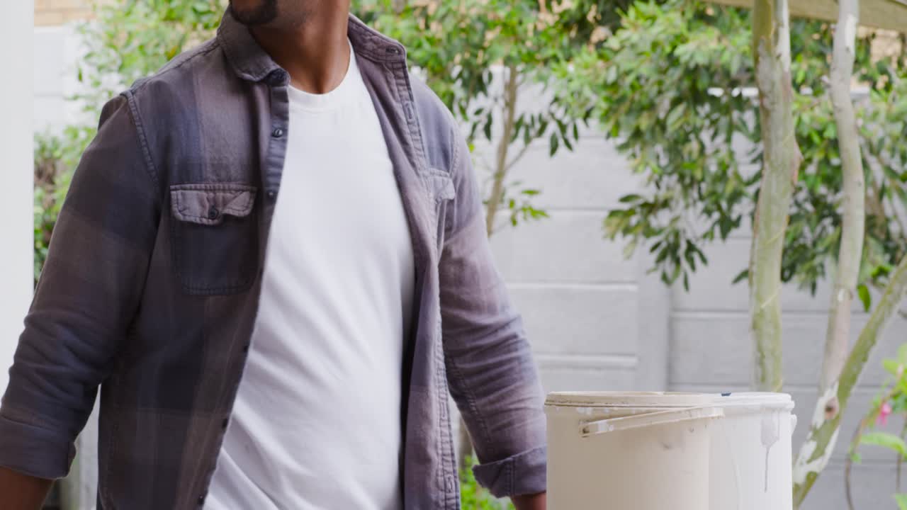 Uomo africano adulto che porta un secchio, lo posiziona su un tavolo con un vassoio, osservando sopra per pianificare la pittura.