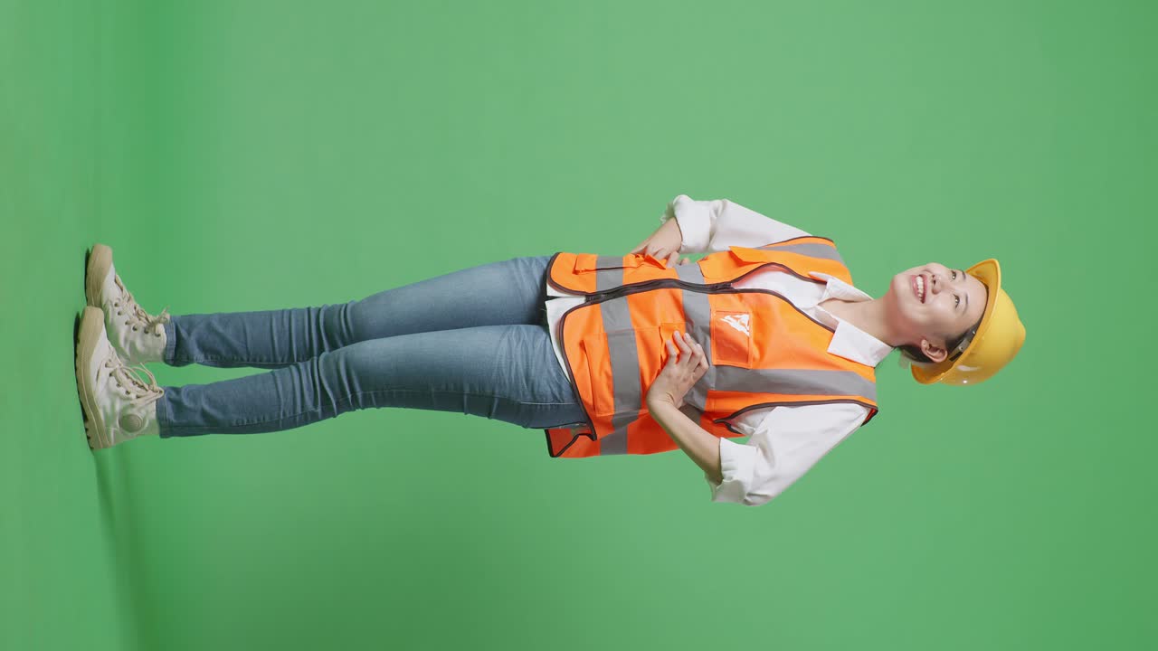 vista lateral completa del cuerpo de una ingeniera asiática con casco de seguridad mirando a su alrededor mientras está de pie con los brazos akimbo en el estudio de fondo de pantalla verde