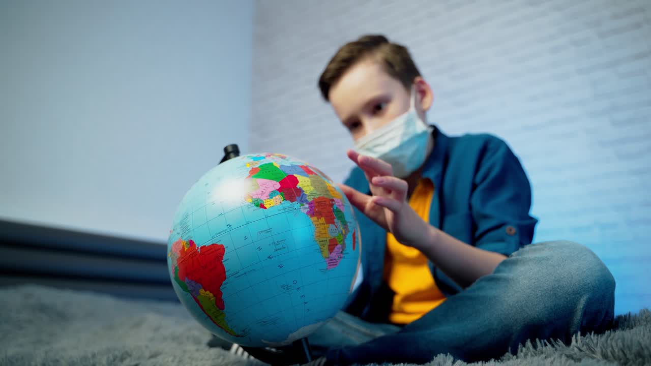 Boy looking at globe learning about virus spreading around the world. Boy wearing medical face mask. Concept of COVID-19 pandemic infection.
