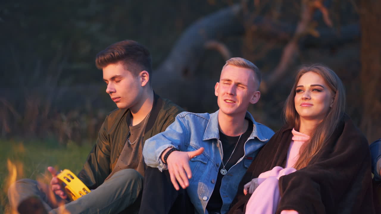 Having good time at picnic in the evening. Friends relaxing, singing songs and playing musical instruments near campfire. Nature blurred backdrop.
