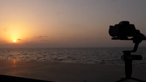 silueta de la cámara en el estabilizador de cardán filmando la puesta de sol en el horizonte sobre el carro de movimiento lento del océano