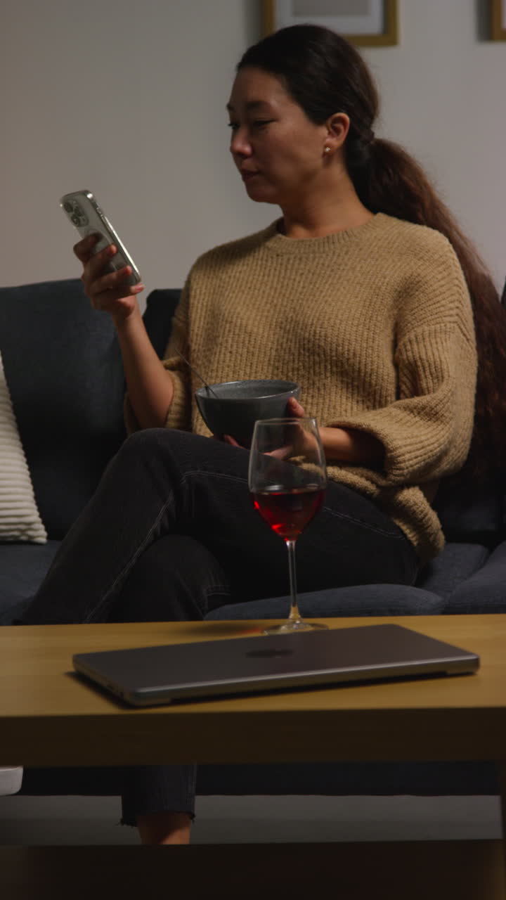 Vertical Video Of Woman At Home Sitting On Sofa Eating Healthy Vegetarian Or Vegan Meal Of Tabbouleh Salad With Glass Of Wine Looking At Mobile Phone