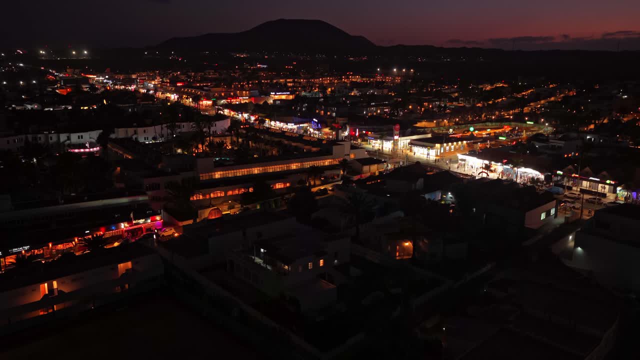 Drone ascends over Corralejo Viejo at night, unveiling a glowing urban grid, beachfront lights, and the dark silhouette of volcanic terrain beyond, blending coastal vibrancy with natural depth