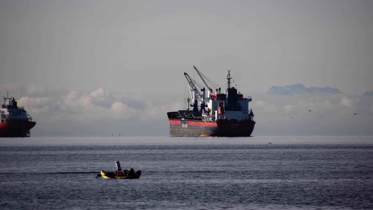 흐린 날 카약을 탄 사람들이 삶을 즐기는 동안 정박지에 데크 크레인이 있는 거대한 빈 벌크선이 배경에서 주문을 기다리고 있습니다.