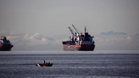 Enorme-Granelero-Vacío-Con-Grúas-De-Cubierta-En-El-Fondeadero-Esperando-órdenes-En-El-Fondo-Mientras-La-Gente-En-Un-Kayak-Disfruta-De-La-Vida-En-Un-Día-Nublado