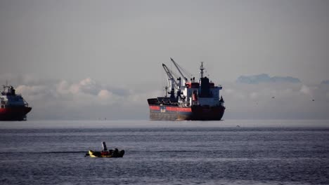 enorme granelero vacío con grúas de cubierta en el fondeadero esperando órdenes en el fondo mientras la gente en un kayak disfruta de la vida en un día nublado